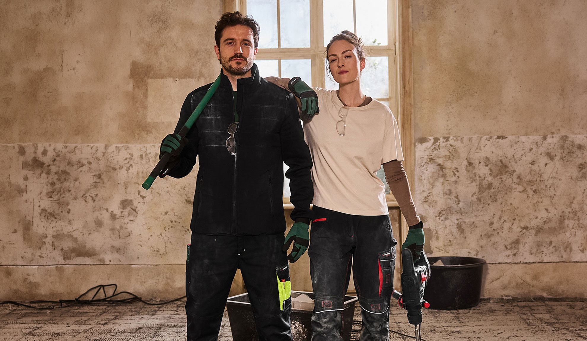 Zwei Bauarbeiter in Arbeitskleidung, Mann mit Schaufel und Frau mit Bohrmaschine.