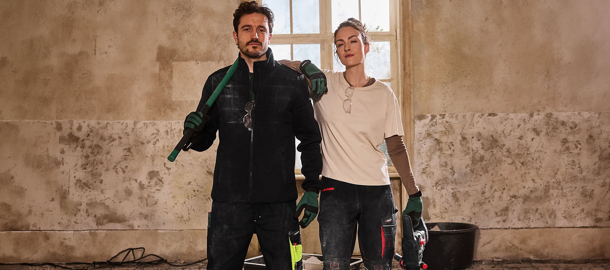 Mann und Frau in Arbeitskleidung, mit Werkzeugen, stehen in einem Renovierungsraum.