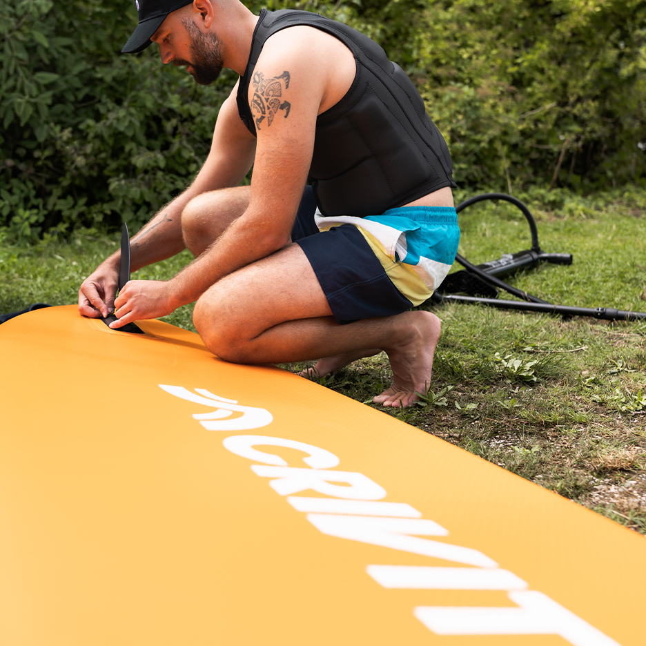 Mann montiert ein orangefarbenes Paddleboard, trägt eine Schwimmweste und Badeshorts.