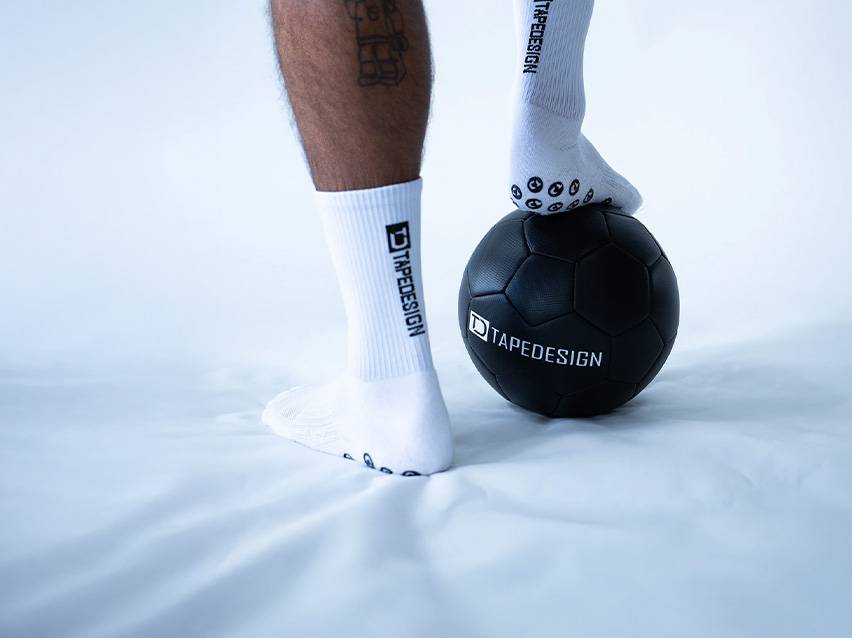 Weiße Sportsocken und ein schwarzer Fußball mit Tapedesign-Logo auf weißem Hintergrund.