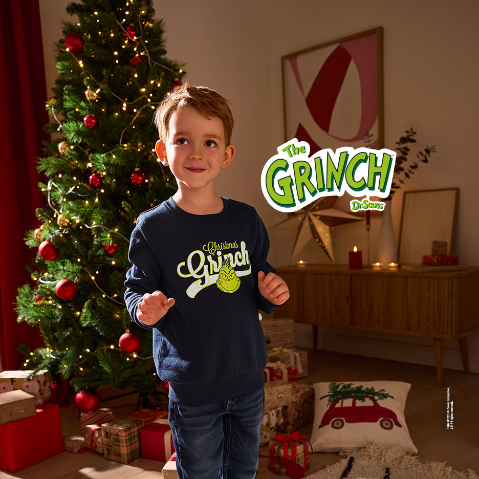 Junge in Grinch-Pullover vor Weihnachtsbaum und Geschenken.