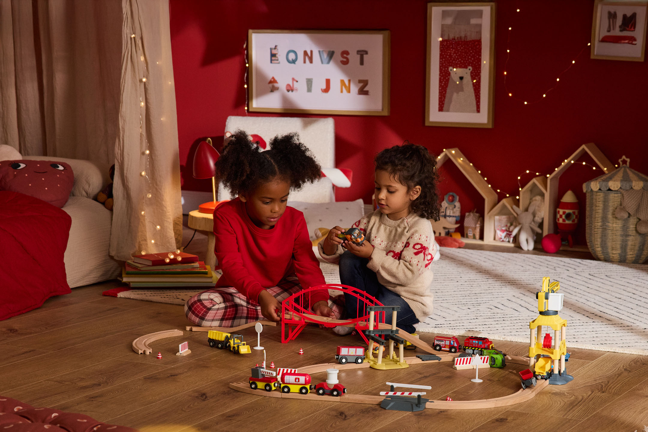 Zwei Kinder spielen mit einer Holzeisenbahn und Bauspielzeug in einem festlich dekorierten Zimmer.