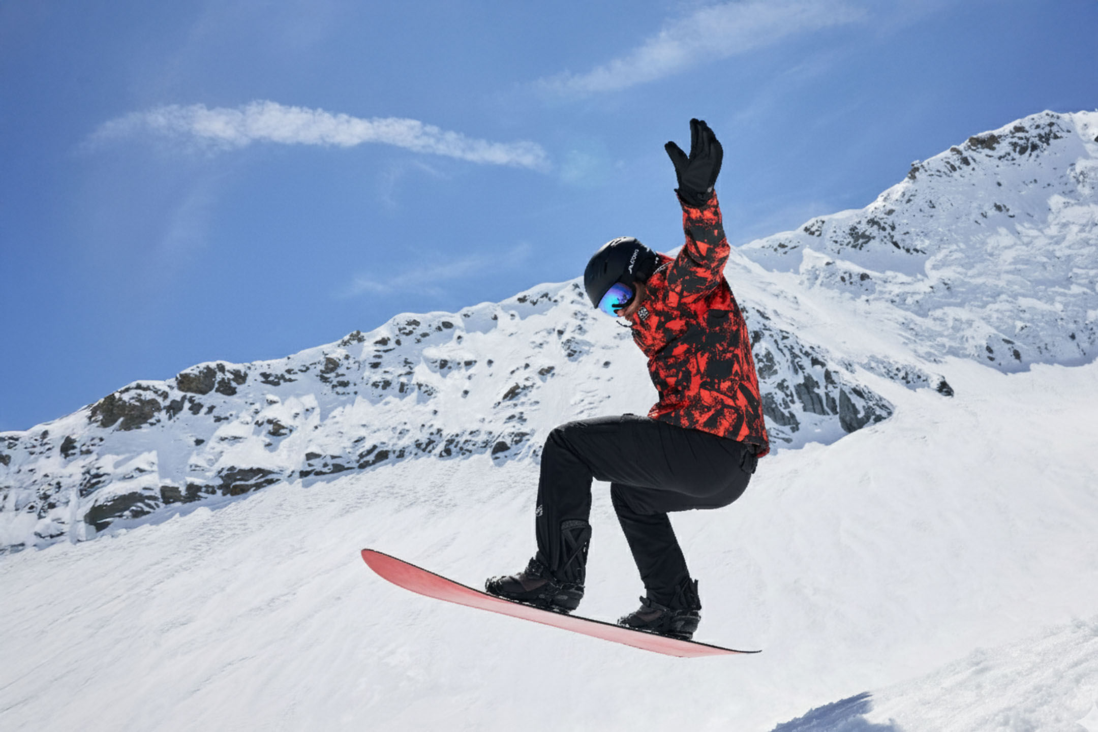 Snowboarder in rot-schwarzer Jacke, schwarzer Hose, Helm und Brille in der Luft auf einem verschneiten Berg.