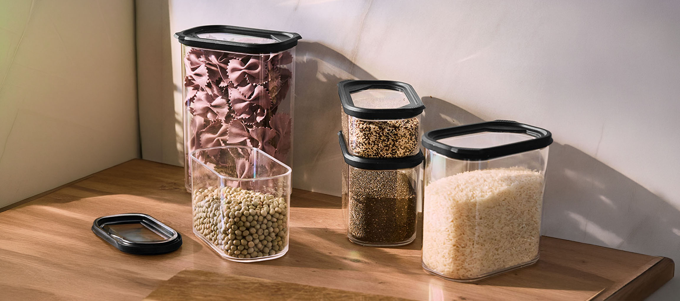 Transparente Küchenbehälter mit schwarzen Deckeln, gefüllt mit Nudeln, Linsen, Quinoa, Chiasamen und Reis auf einer Holzoberfläche.