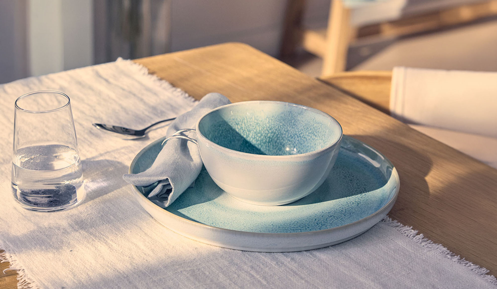 Gedeckter Tisch mit blauer Schale, Teller, Wasserglas und Serviette auf einem Holztisch.