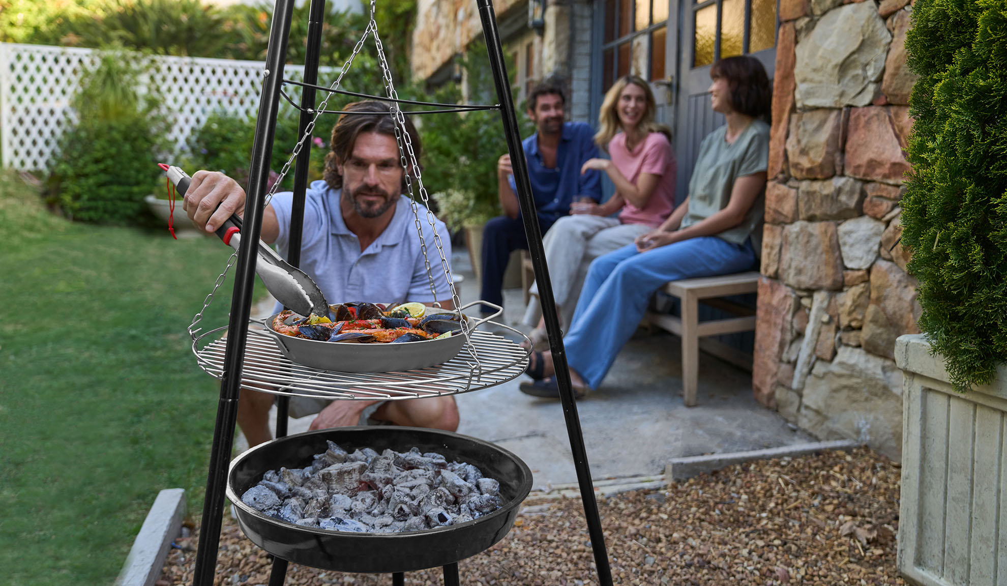 Mann grillt Meeresfrüchte auf Dreibein, Freunde entspannen im Hintergrund.
