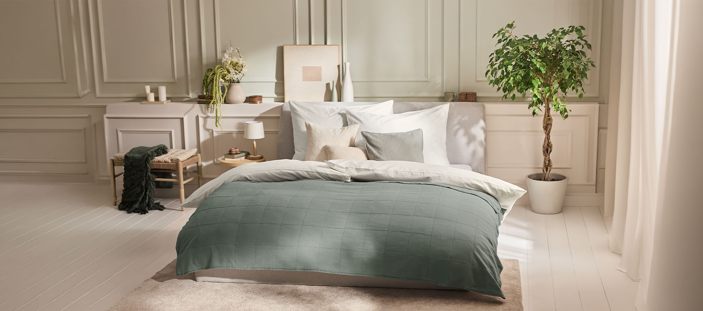 Schlafzimmer mit Bett, grüner Bettdecke, Kissen, Nachttisch und einer Pflanze.