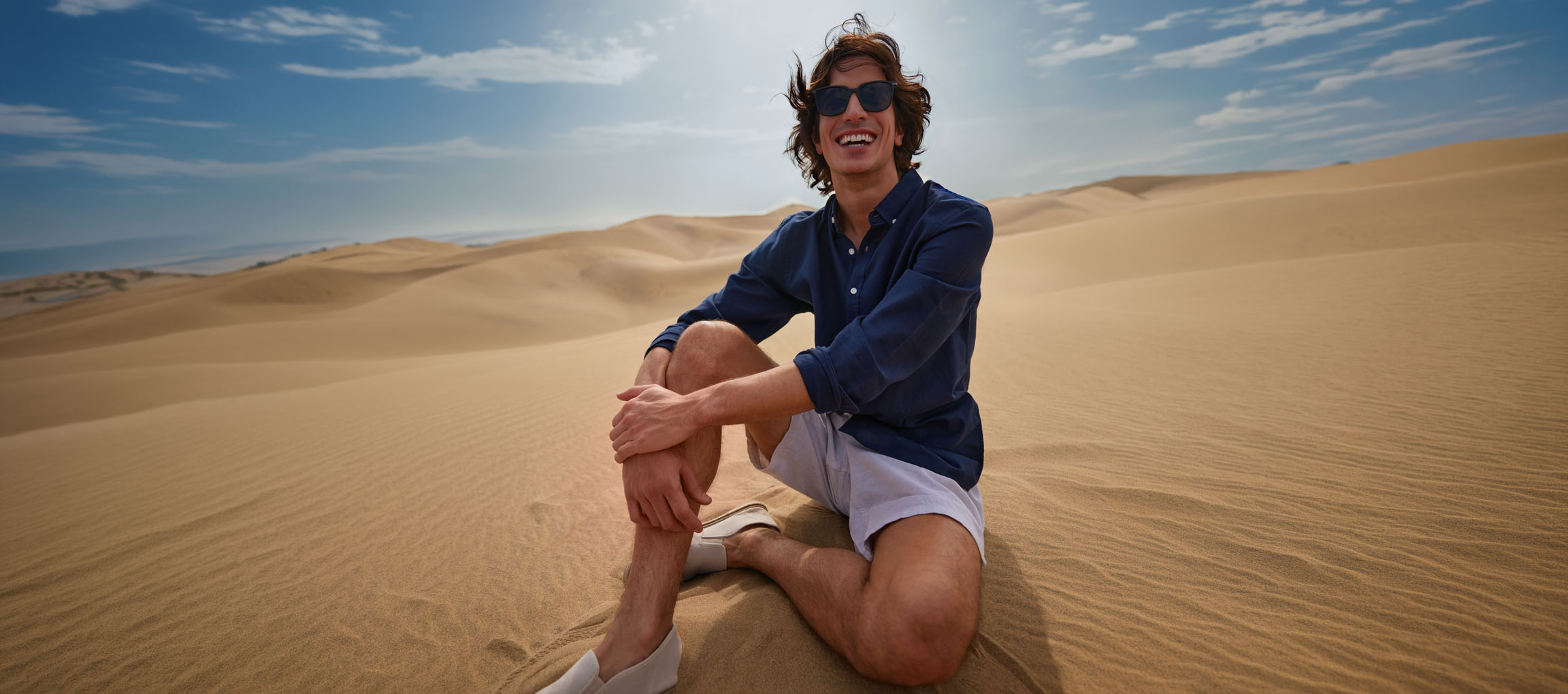 Mann in dunkelblauem Hemd und Shorts sitzt lächelnd in Sanddünen.