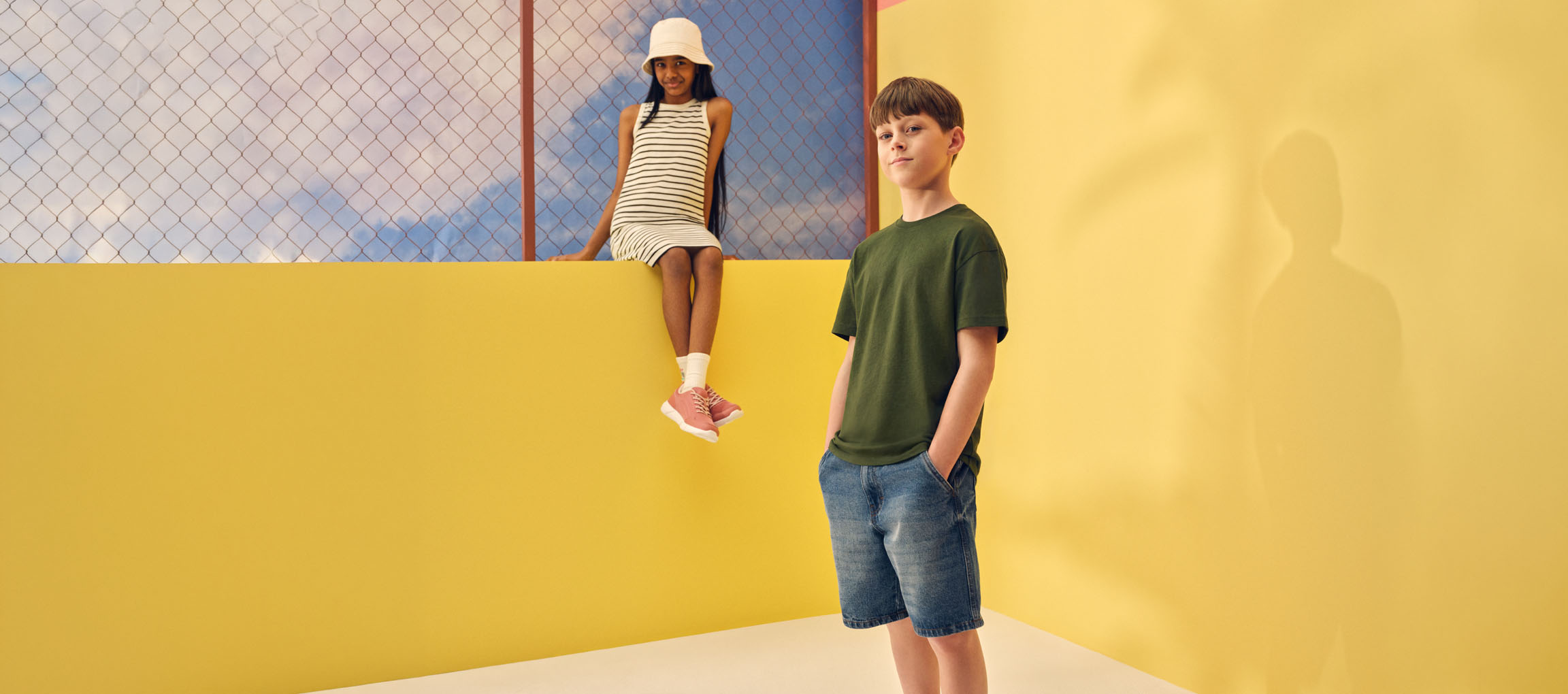 Zwei Kinder in lässiger Sommerkleidung: ein Mädchen im Ringelkleid und ein Junge im grünen T-Shirt und Jeansshorts.