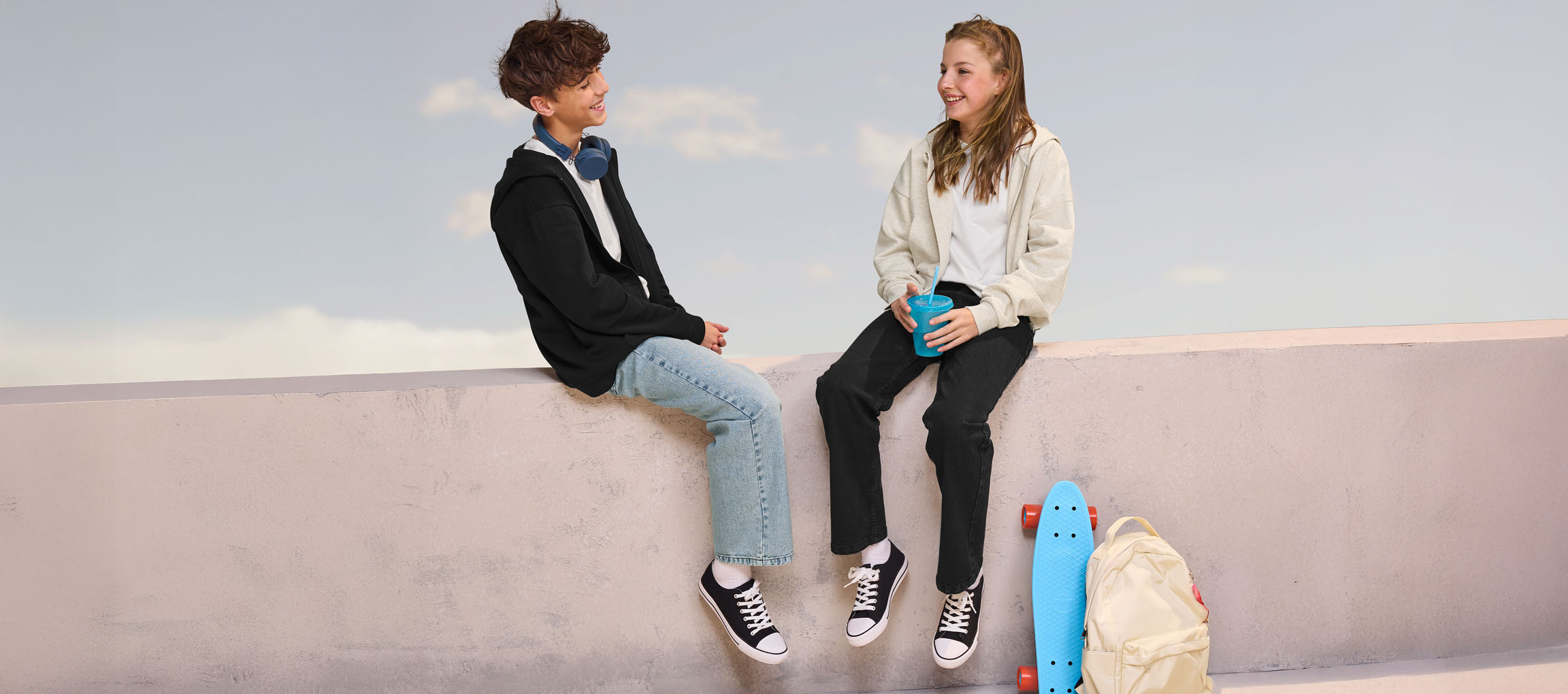 Zwei Teenager in Hoodies und Jeans sitzen auf einer Mauer, mit Skateboard und Rucksack in der Nähe.