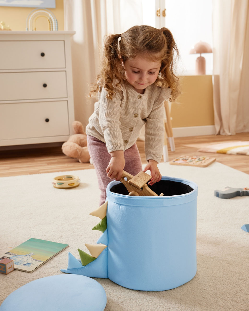 Ein Kind legt Holzspielzeug in eine blaue Aufbewahrungsbox mit Dinosaurier-Motiv.