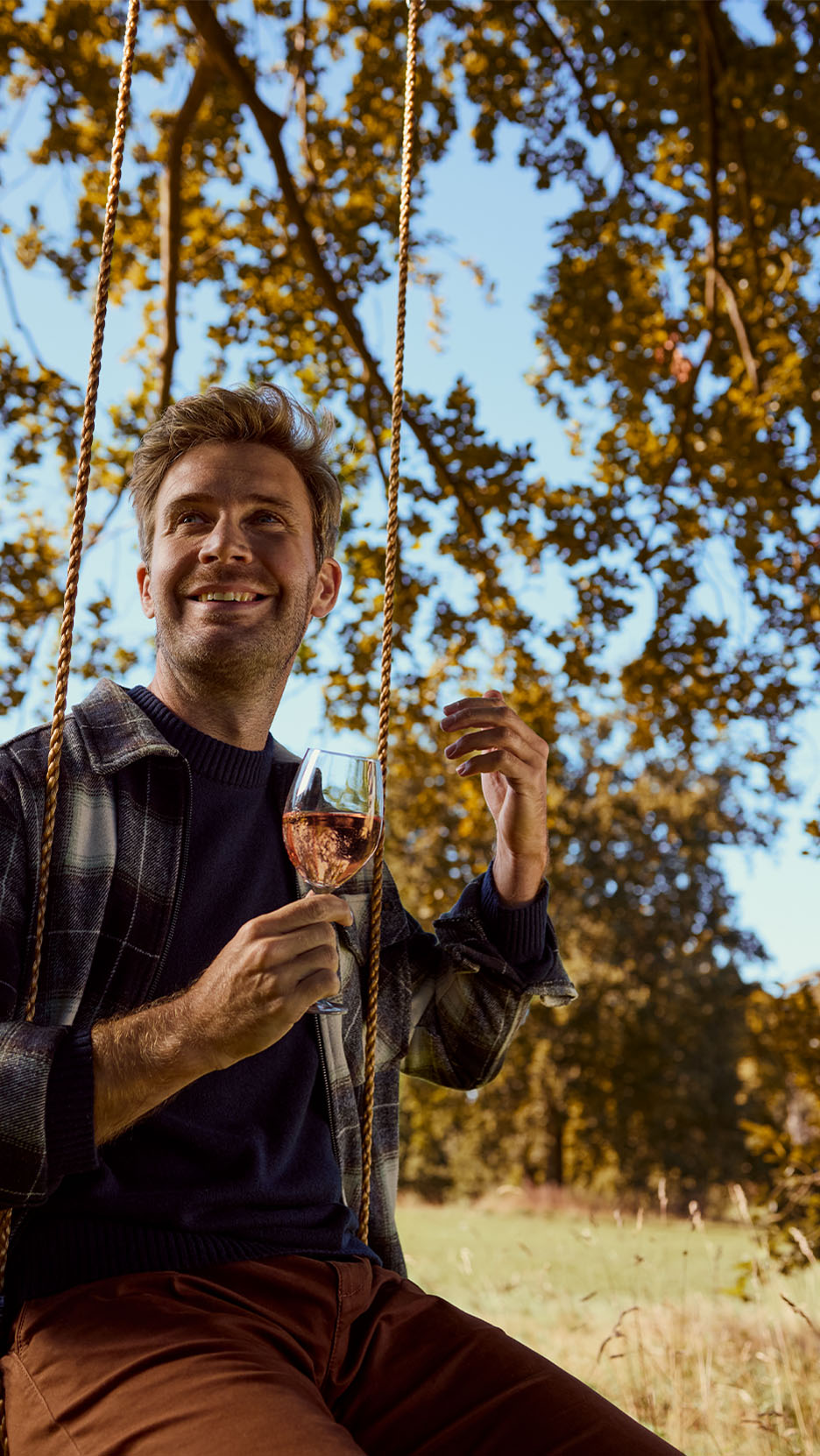 Ein Mann auf einer Schaukel trinkt Roséwein, gekleidet in ein kariertes Hemd und einen dunkelblauen Pullover.