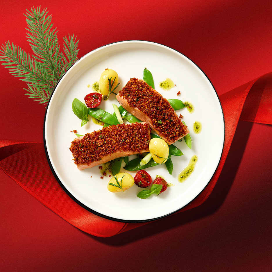 Lachsfilets mit knuspriger Kruste, neuen Kartoffeln, Zuckerschoten und Kirschtomaten auf einem weißen Teller, mit festlichem rotem Hintergrund und Tannenzweig.