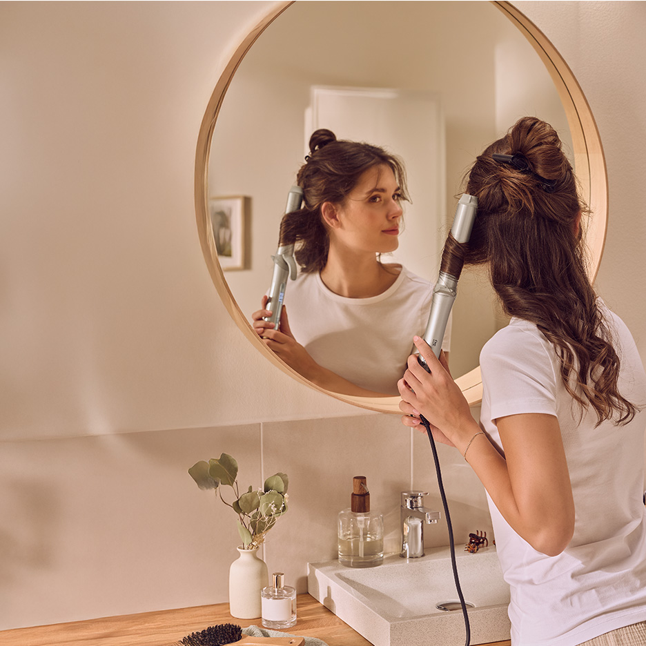 Frau lockt sich die Haare mit einem Lockenstab vor einem runden Spiegel im Badezimmer.