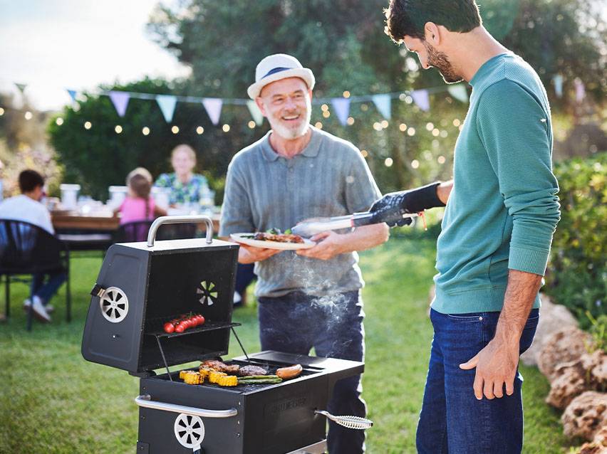 Zwei Männer grillen im Garten, einer hält einen Teller mit Grillgut, der andere bedient den Grill.