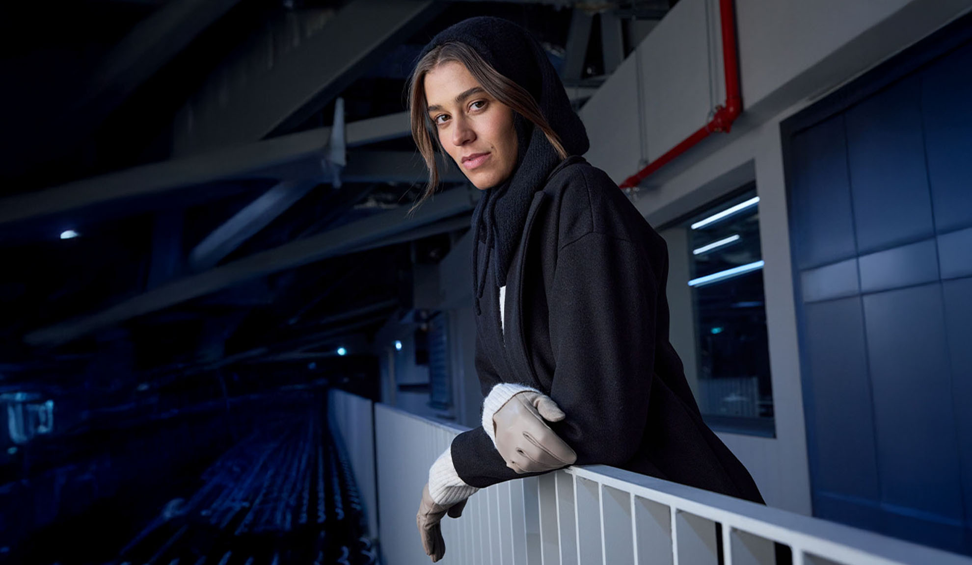 Frau in schwarzem Mantel und Kapuzenschal, an einem Geländer in moderner Umgebung lehnend.