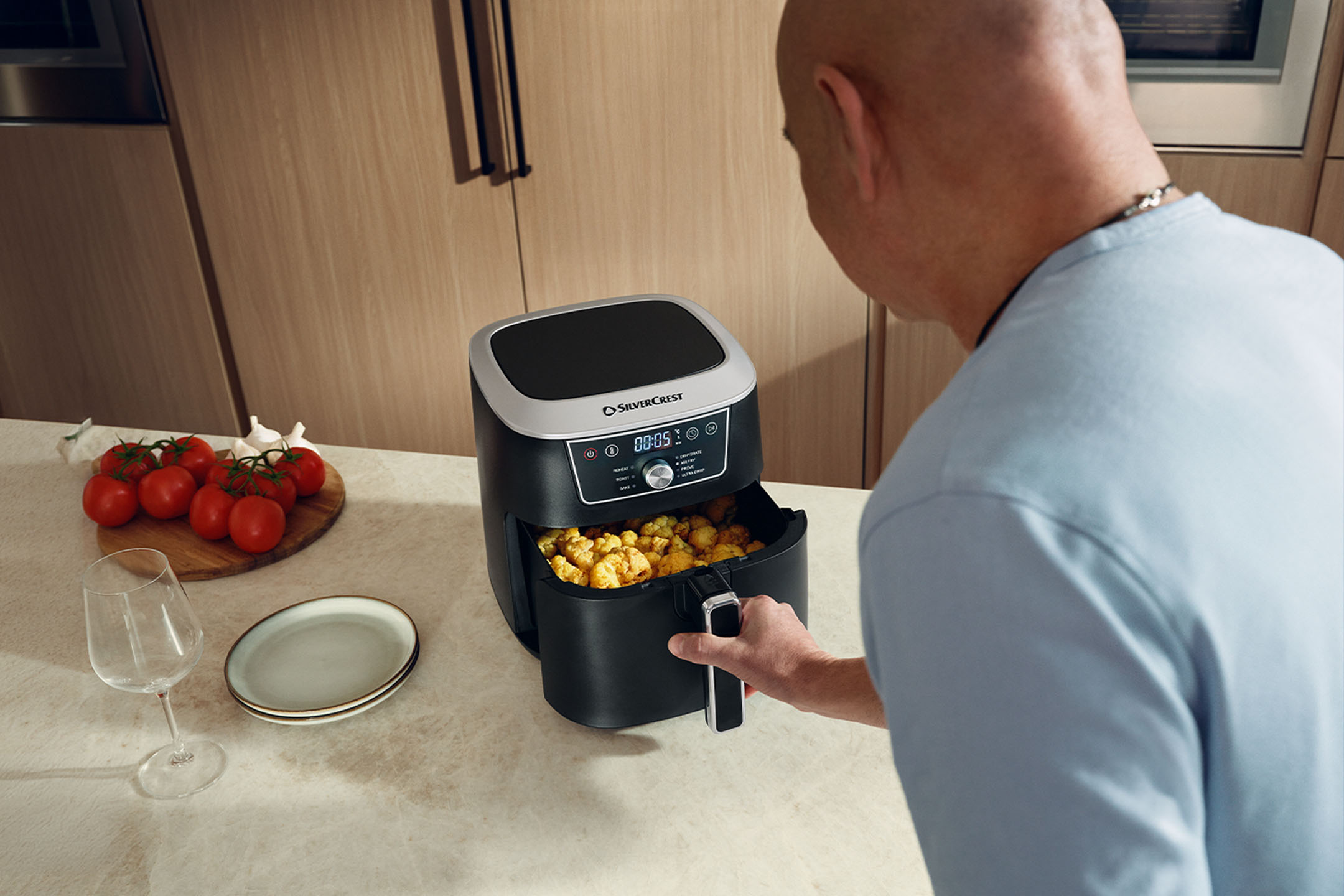 Mann kocht mit einer Heißluftfritteuse in der Küche, mit Tomaten und Tellern.