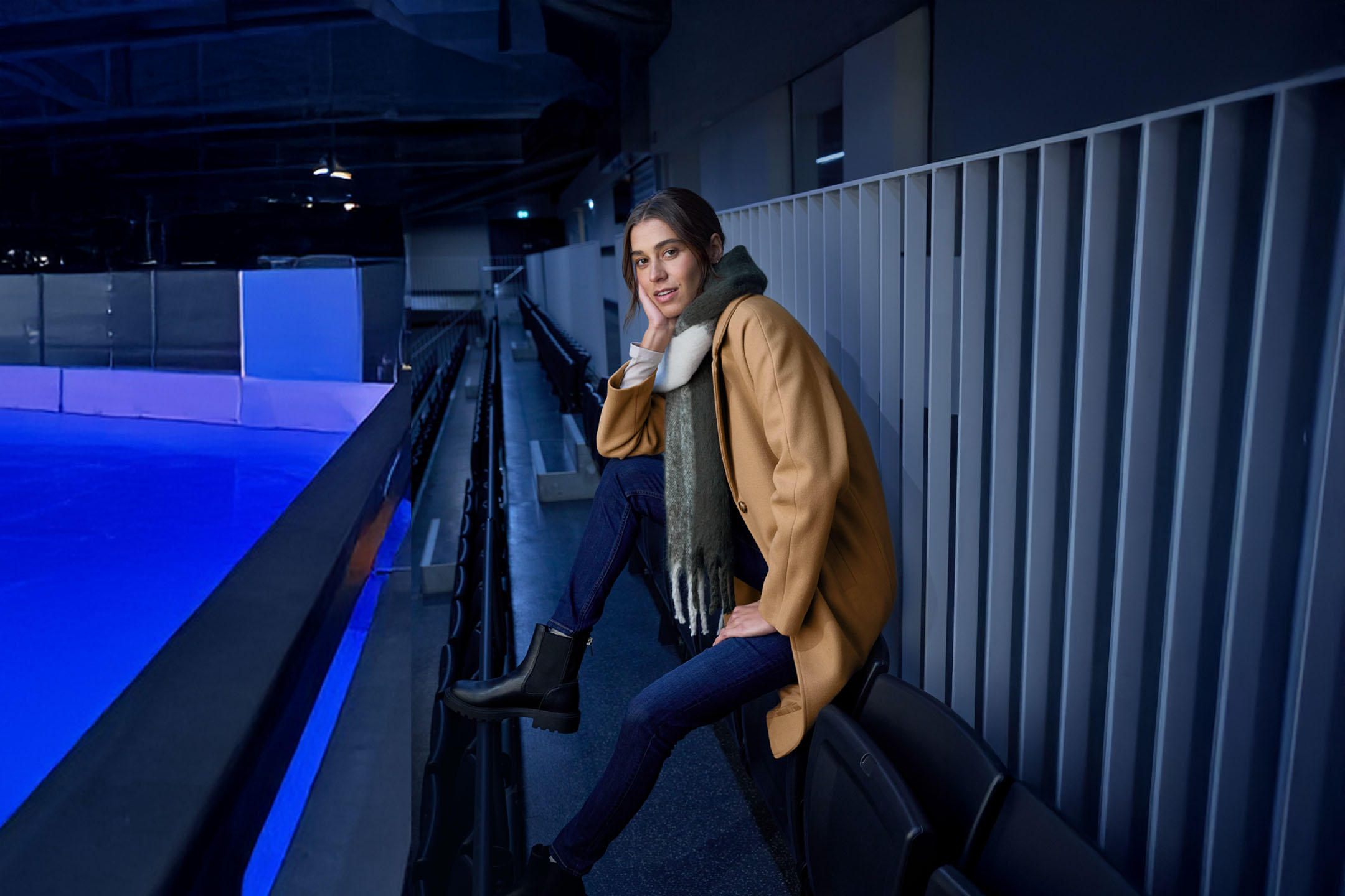 Frau in Mantel und Schal sitzt auf der Tribüne einer Eisbahn.