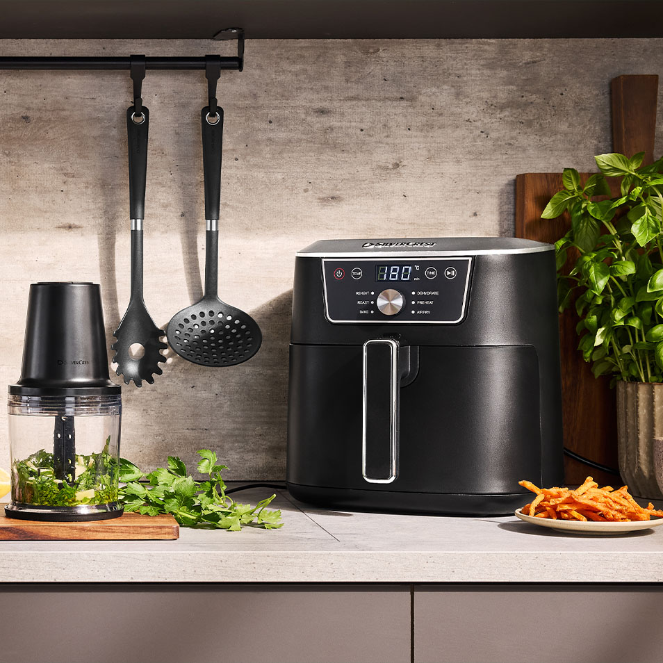 SilverCrest Heißluftfritteuse und Zerkleinerer auf einer Küchenarbeitsplatte mit frischen Kräutern und Süßkartoffelpommes.