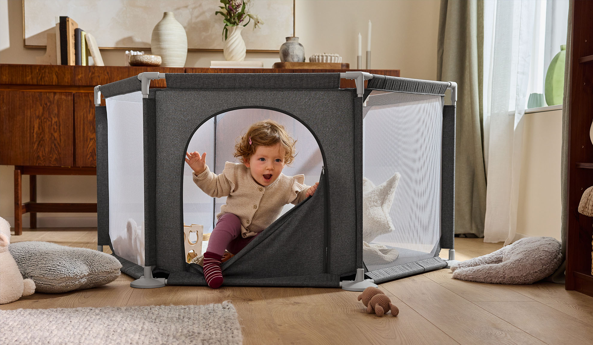 Glückliches Baby krabbelt aus einem grauen Laufstall im gemütlichen Wohnzimmer.