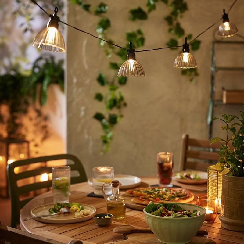 Außenbeleuchtung mit warmen Lichtern über einem Holztisch mit Essen.